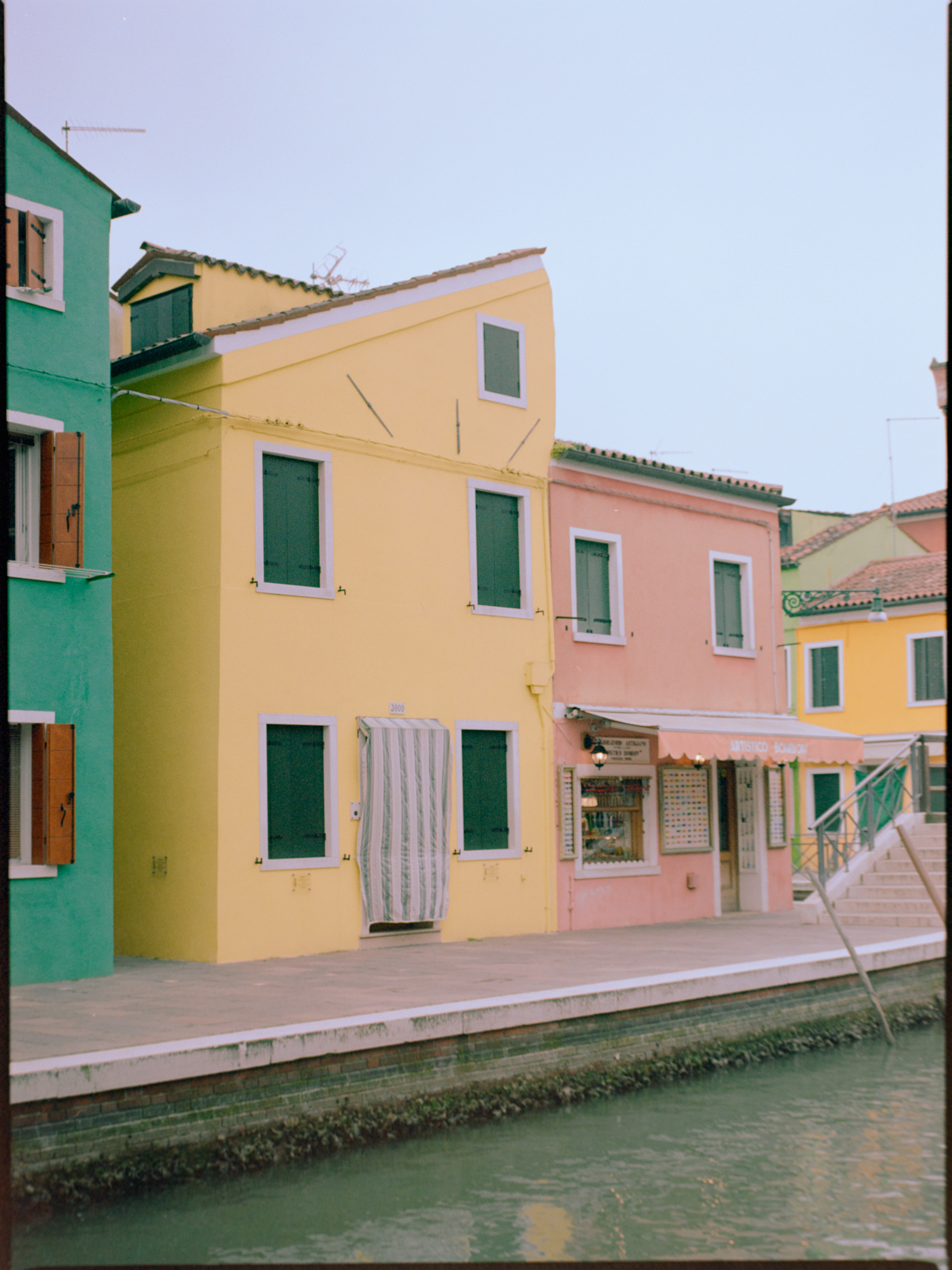 Row of brightly painted houses along a Burano canal with green shutters — Venice on Kodak Gold 200