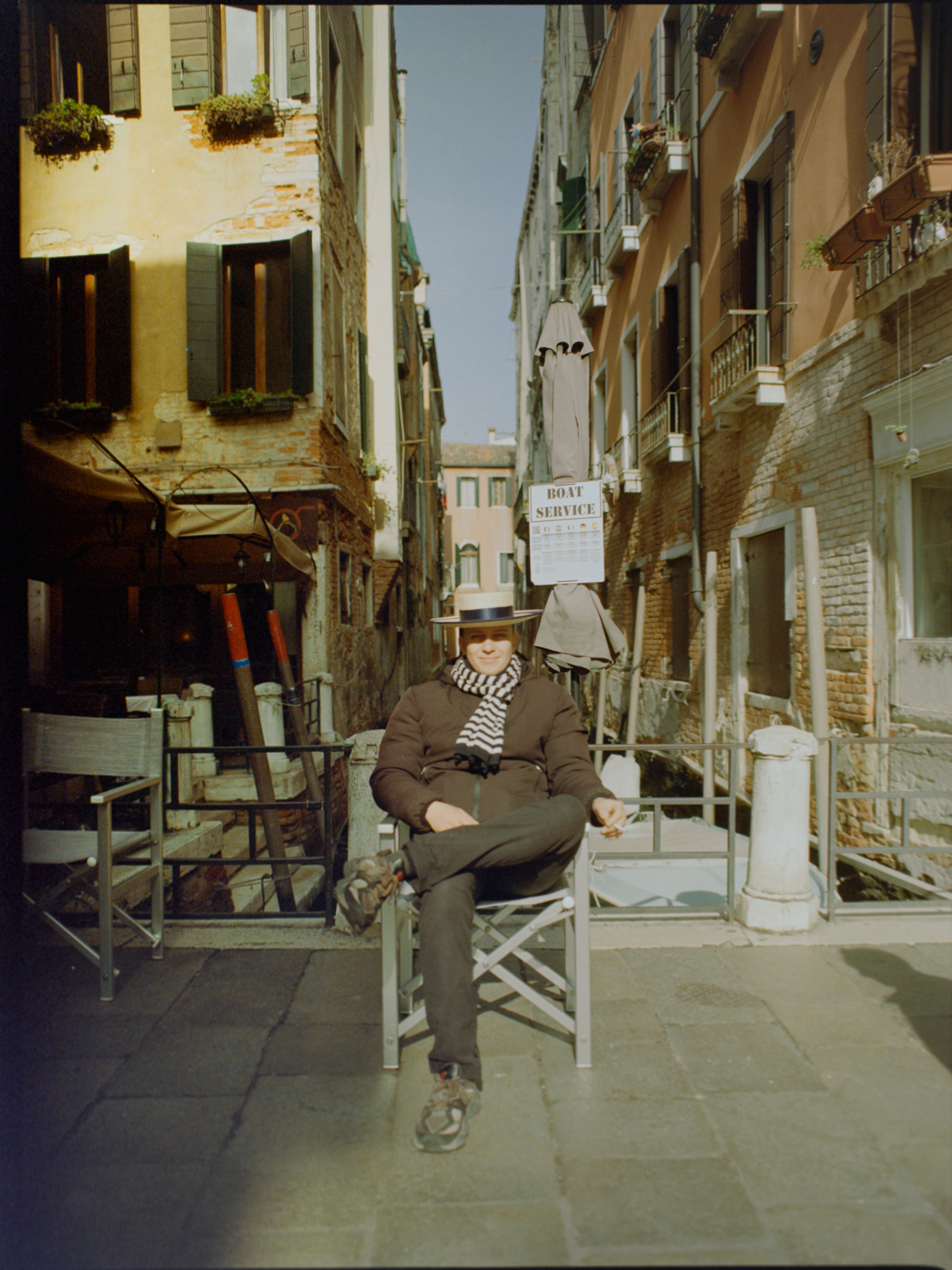 Gondolier in traditional dress seated by a canal, arms crossed, waiting for passengers — Venice portrait on medium format film