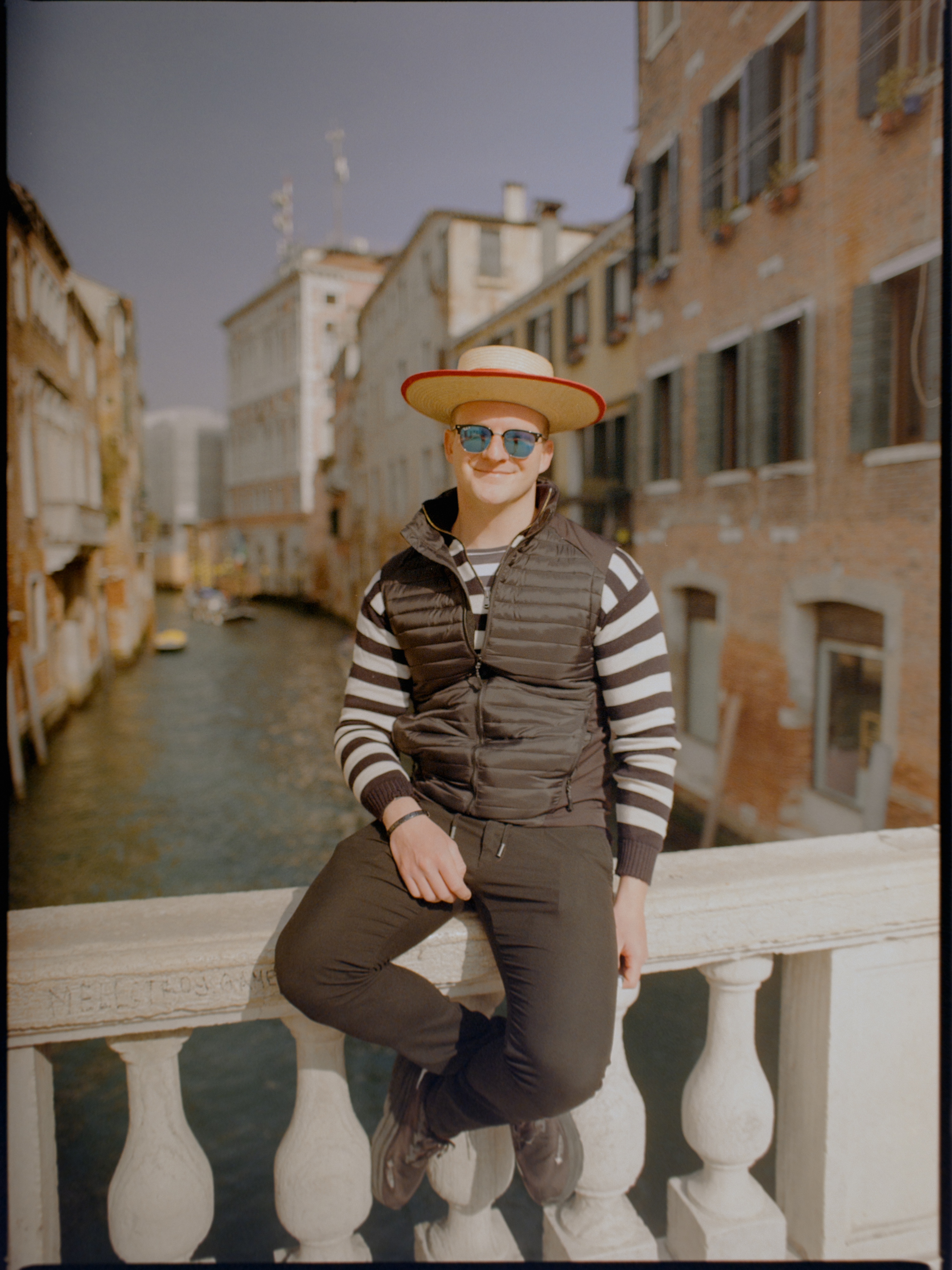Young gondolier in striped shirt and straw hat sitting on a bridge railing — Venice portrait on film
