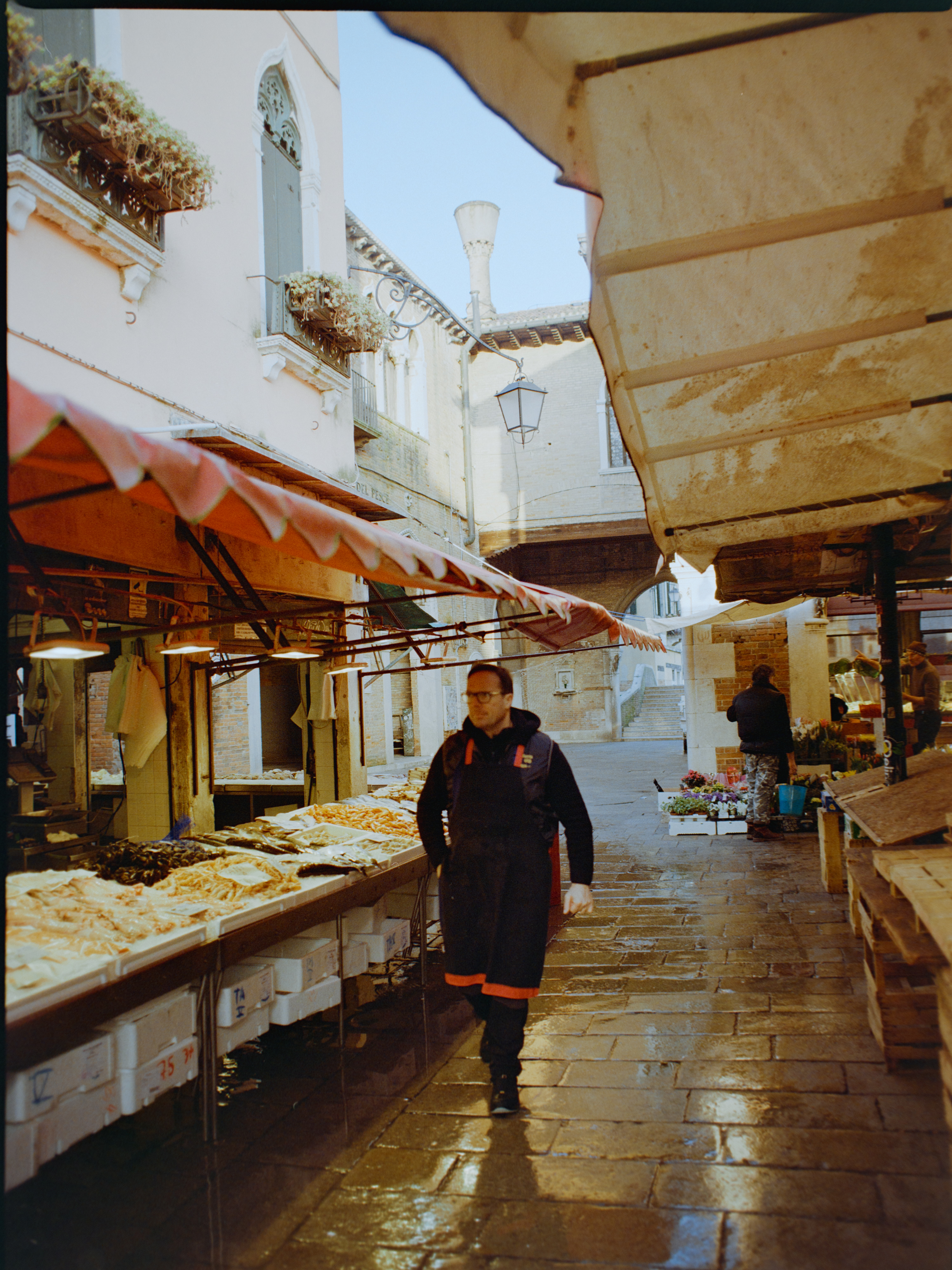 Wet narrow aisle between fish market stalls with red awnings and single vendor — Venice Rialto Market on film